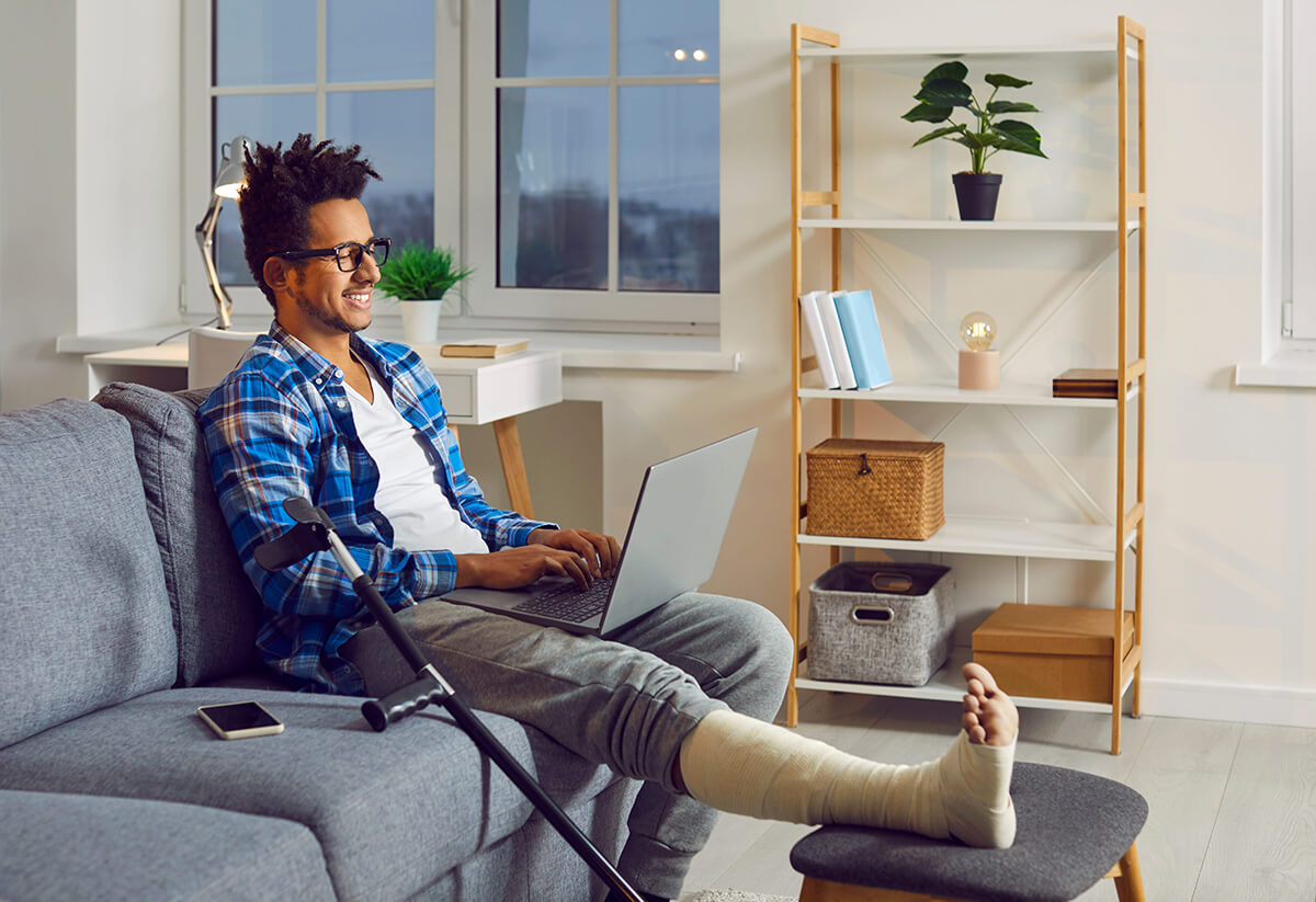 Man recovering at home with a leg injury while using a laptop, representing financial support and recovery after an accident.